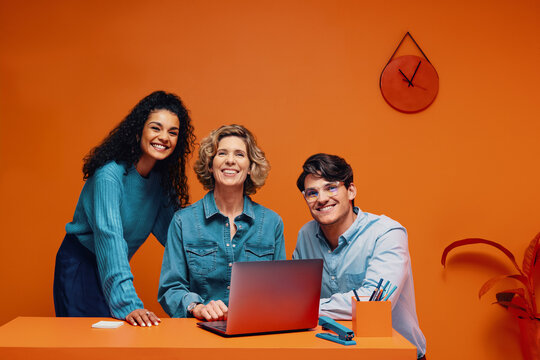 Diverse team of co-workers happily posing in vibrant office with laptop and colourful backgrounds
