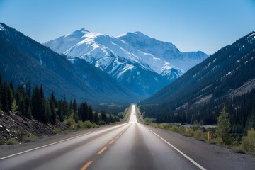 Fototapeta premium Straight road flanked by coniferous forests and snow capped mountains, sunny blue sky