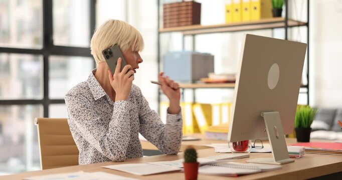 Smiling young business woman talking on phone sitting at office desk. Professional secretary holding mobile phone and consulting with client
