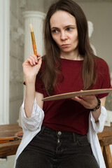 Artist looks thoughtfully at drawing canvas while painting and holding palette and brush in hands