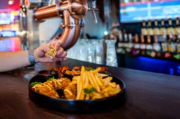 hand picking up a piece of food from a platter with fried chicken wings, French fries, and dipping sauces, set against blurred bar background. Ideal for themes of dining, food, and social gatherings.