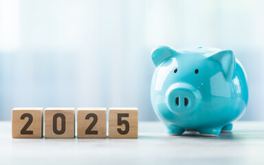 2025 is written on a wooden block beside a A blue piggy bank on a wooden table against a bright white background. Money planning and Financial planning, savings, and wise money management.