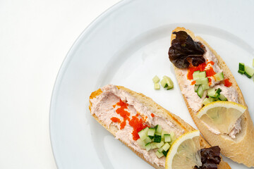 Tasty open-faced sandwiches with tuna spread, topped with fresh vegetables and lemon slices on a white plate
