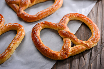 A close-up shot of freshly baked, golden-brown pretzels resting on parchment paper. The pretzels have a glossy, and the wooden background adds a rustic charm to this appetizing scene