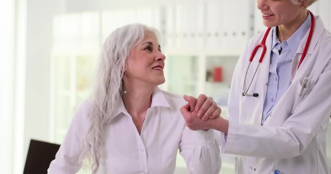 Elderly woman visits doctor and receives optimistic news after medical examination. Therapist holding hand of old patient and giving hope