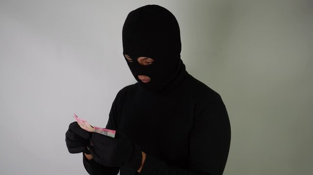 4k video footage of Dangerous young Adult black masked robber is holding and counting a bunch of indonesian paper money bills after stealing from his victim.