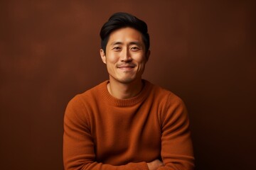 Fototapeta premium Portrait of a content asian man in his 30s wearing a cozy sweater while standing against soft brown background