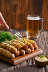 Sausages grilled to perfection, drizzled with mustard and mayonnaise, served on a wooden board alongside fresh parsley and a mug of beer. A hand with a fork is ready to dig into this delicious feast