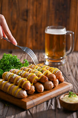 Sausages grilled to perfection, drizzled with mustard and mayonnaise, served on a wooden board alongside fresh parsley and a mug of beer. A hand with a fork is ready to dig into this delicious feast