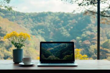 An image of a laptop on a wooden table with a white cup and a small vase of yellow flowers next to it. The background shows a view of lush green mountains through a large window.