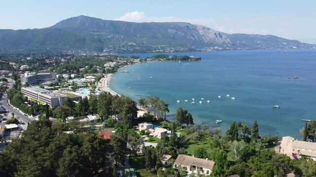 Dolly in aerial flight over Dassia Bay and Beach during daytime, overlooking the sea, coastline, and mountains in Corfu, Greece