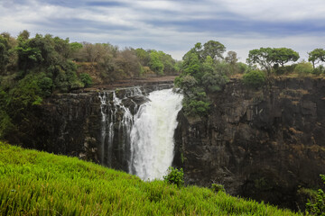 Water cascades powerfully over Victoria Falls during a cloudy day in Zimbabwe, surrounded by lush...