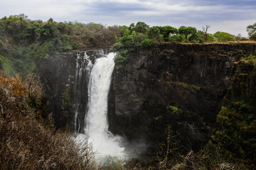 Victoria Falls roars in Zimbabwe, showcasing its majestic cascade amidst lush greenery on a cloudy...