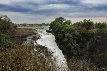 Water cascades over rocky cliffs at Victoria Falls in Zimbabwe, surrounded by lush greenery and...