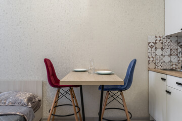 small simple kitchen in the apartment. White kitchen furniture, refrigerator, microwave, soft chairs in blue and red colors. Affordable real estate. There are plates on the table. Cozy dinner at home