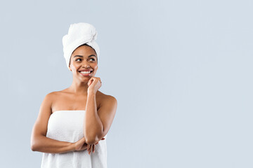 Cool offer in spa. Excited young african woman wrapped in white bath towels looking at copy space, grey studio background