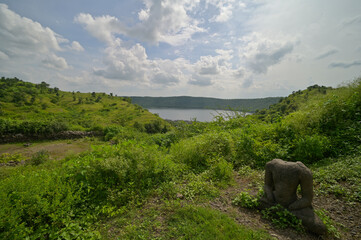 Ancient headless stone sculpture in meditative seating posture amidst greenery near Lonar crater,...