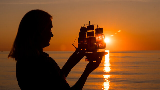 A young woman holds a toy boat in her hands. Standing at sunset by the sea