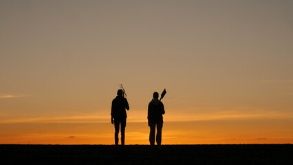 In a field at sunset, silhouettes of a couple of farmers with working equipment can be observed from the back.