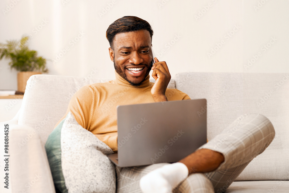Aufkleber Smiling Afro Freelancer Guy Working On Laptop Computer And Chatting On Phone Sitting On Sofa At Home. Empty Space