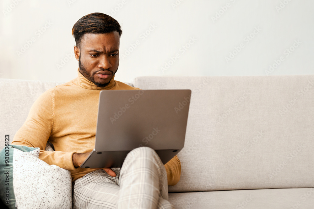 Poster serious afro businessman working on laptop computer sitting on sofa at home. copy space, selective f