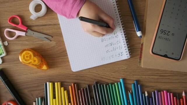 Child doing math test in notebook using smartphone calculator to solve equation with stationery on wooden table. Girl hand writing mathematical exercises as school homework. Top view