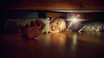 Emotional little boy, child lying on the floor and looking under bed with phone flashlight,...