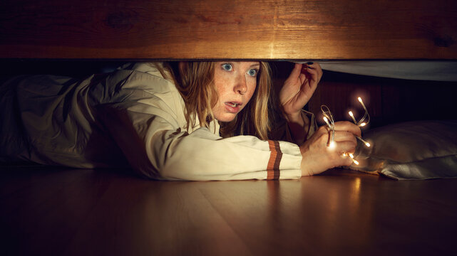 Young woman with emotional face, holding lights, lying on floor and looking under bed with emotions of fear. Spooky season. Trick and treat. Concept of human emotions, holiday