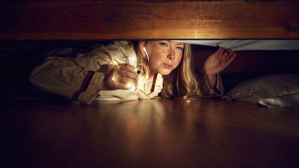 Young emotive woman lying on floor and looking under bed with light, looking with careful, fearful gaze. Concept of human emotions, lifestyle, holiday, secrets, spooky vibe