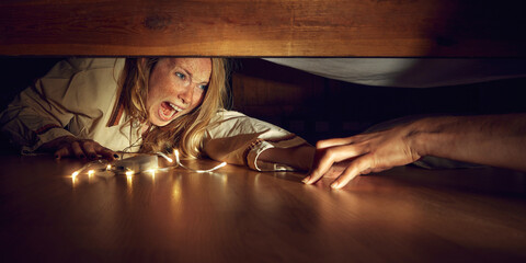 Young fearful woman lying under bed with light while male hand grabbing her. Woman shouting in fear and horror. Spooky season. Concept of Halloween, human emotions