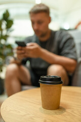 Young man with phone and power bank. Takeaway paper coffee cup with aroma latte coffee on a wood table at city cafe. Male with social media addiction on smartphone scrolling internet, 