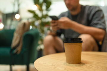 Young man with phone and power bank. Takeaway paper coffee cup with aroma latte coffee on a wood table at city cafe. Male with social media addiction on smartphone scrolling internet, 