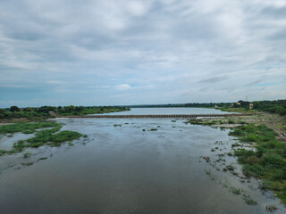 A serene countryside river flows under a bridge, reflecting a blue sky dotted with white clouds. Clear water laps at grassy banks, where a small temple sits peacefully, embraced by nature’s beauty.