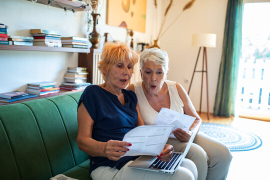 Senior lesbian couple holding bill paperwork and using laptop at home