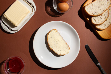 Toast with butter and sweet jam on the white plate. Flat lay of the breakfast preparation on brown background. Traditional breakfast concept. Top view