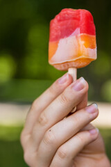 Closeup of the hand holding colorful refreshing partially melted bitten fruit ice cream popsicle on a hot summer day, Selective focus, Burred green outdoor background. Copy space.

