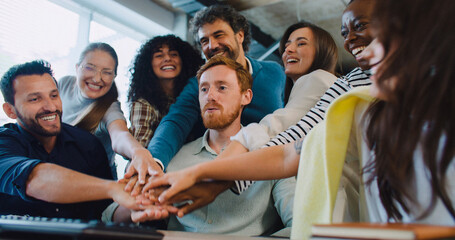 Camera focus of smart Caucasian male sitting in middle of his working team. Everyone smiling with joy after successful team building process. Raising their hands. Keyboard visible on desk. Office room