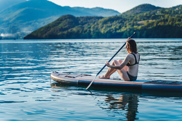 Fototapeta premium Young cheerful woman sitting with paddle on sup surf board on water in mountain lake