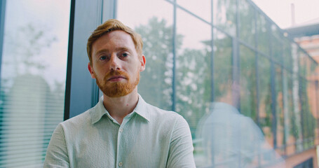 Handsome Caucasian man with red hair and beard standing in front of large glass window. Modern...