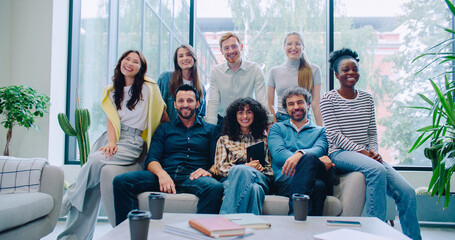 Camera moving closer to multi-ethnic group of friends. Everyone gathering together next to one sofa in lounge area. Engineers or programmers posing for camera in big room. Sunny weather in background.