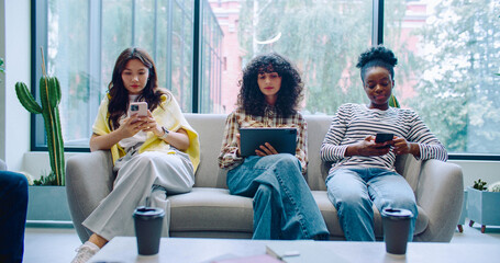 Camera view of three girls sitting on couch on big resting area. Using gadgets. Scrolling information or social media on smartphones and tablet. Catching up on news during coffee break.