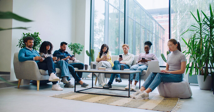 Large room in modern building. Lounge area. People sitting on couches while waiting in line. Woman with stack of paper passing by. Everyone using smartphones or tablets. Girl with laptop. Antisocial.
