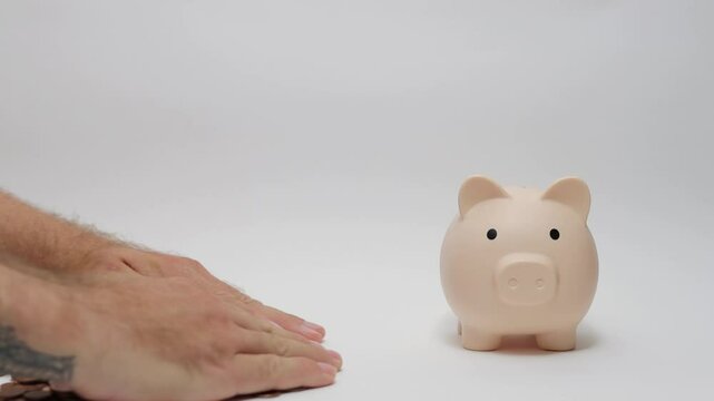 Man pushes scattered coins toward piggy bank on white surface. Coins are spread out, and hands are used to gather them efficiently