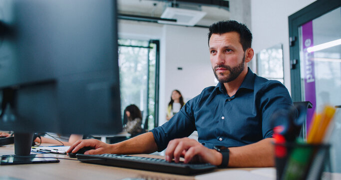 Camera view of gorgeous Caucasian man in shirt working in business company. Busy specialist looking directly at screen and using keyboard to type message. In background people sitting at computers.