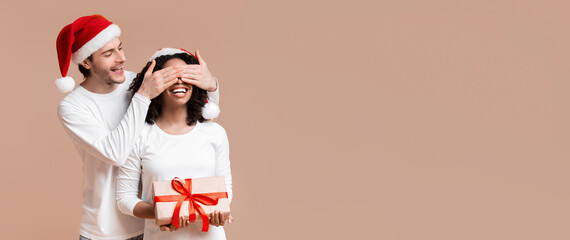 Personal Santa. Loving Boyfriend Making Christmas Surprise to His Black Girlfriend, Covering Her Eyes And Presenting Gift, Yellow Studio Background