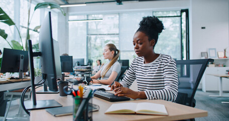 Portrait African American woman with casual clothes working at computer in office. Distracted by camera from work and joyfully smiling. In background people sitting at their workplaces. Job concept.