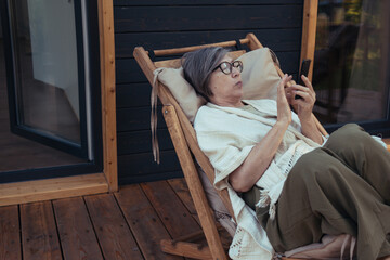 Senior asian woman sitting in chair on terrace of wooden house and scrolling news using phone at summer morning