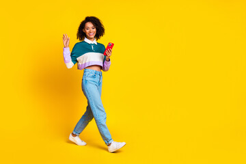 Full length photo of friendly girl wear striped shirt hold smartphone go empty space waving palm isolated on vivid yellow color background