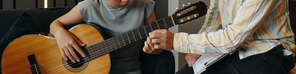Child learning guitar playing skills while teacher guides hand placement, concentrating on...