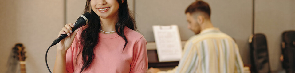 Fototapeta premium Young woman holding microphone and singing in music studio, with musician playing piano in background, both deeply involved in their music and performance